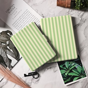 Può includere: Due copertine di libri a righe verdi e crema sono esposte su una superficie di marmo. Le copertine sono rettangolari e presentano un motivo a righe verticali. Sono visibili anche un apribottiglie nero e un libro con testo.