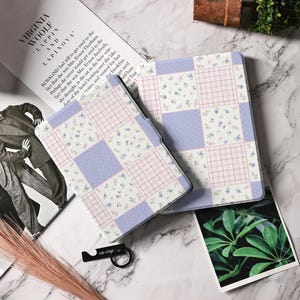 以下が含まれることがあります： パッチワークデザインのタブレットカバー2つ。カバーは、薄紫、ピンクの格子柄、花柄の正方形が特徴です。黒い栓抜きと本も写っています。