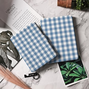 Puede incluir: Dos fundas para tablet con estampado de cuadros vichy azul y blanco se muestran sobre una superficie de mármol. Las fundas están abiertas, revelando un interior negro. También se ve un abridor de botellas negro y hierba seca.