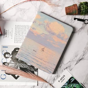 Pode incluir: Uma capa retangular para tablet com uma pintura de paisagem marinha, apresentando um veleiro sob um céu pastel. A capa é feita de um material liso. A estética geral é suave e artística.