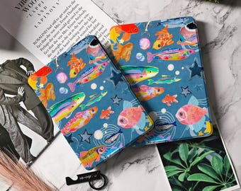 May include: Two blue decorative cases with a colourful fish pattern. The cases feature various fish in shades of orange, pink, yellow, and green, along with white bubbles and blue stars. The cases are placed on a white surface with a book and a plant.