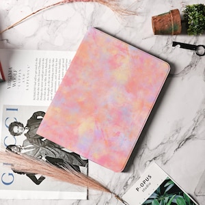 Puede incluir: Una funda rectangular para tableta con un diseño de acuarela en tonos rosa, amarillo y morado. La funda está sobre una superficie de mármol junto a una revista y pequeños objetos decorativos.