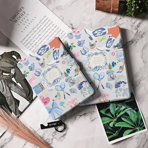 May include: Two light-coloured book covers with a repeating pattern of blue and pink illustrations, including boots, flowers, and a woman's portrait. The text "FOR WE ARE GOD'S MASTERPIECES" is visible. A black bottle opener is in the foreground.