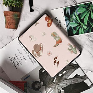 May include: A pink e-reader case with a cowboy theme. The case features a variety of designs, including cowboy hats, boots, cacti, and flowers. The case is made of a durable material and has a sleek design.
