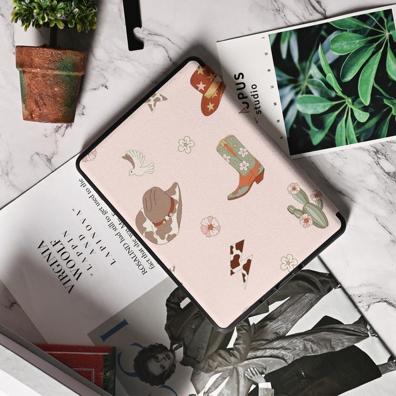 May include: A pink e-reader case with a cowboy theme. The case features a variety of designs, including cowboy hats, boots, cacti, and flowers. The case is made of a durable material and has a sleek design.