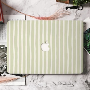 May include: A pale green laptop case featuring a white, vertical striped design. The case has a cut-out for the Apple logo. The case is on a marble surface with other items in the background.