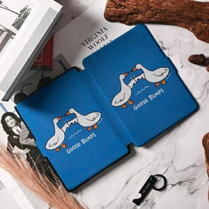 May include: Blue tablet case featuring a cartoon design of two white geese with orange feet and beaks, standing on a wave. The text "Goose Bumps" is below the geese. The case is open, revealing the interior.