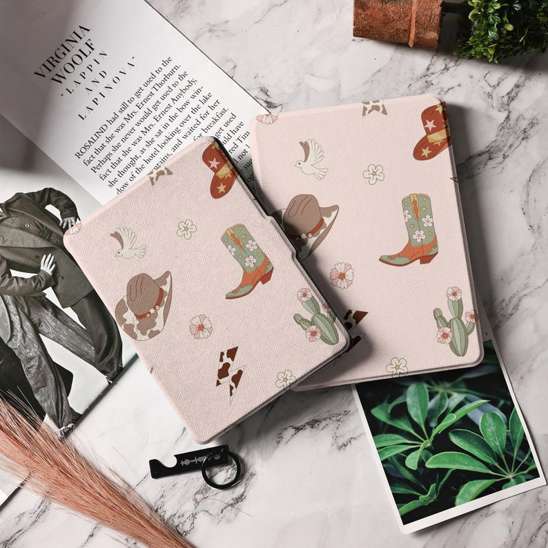 May include: A pink and white Kindle cover with a western theme. The cover features a repeating pattern of cowboy boots, cacti, hats, and birds. The cover is made of a soft, textured material.