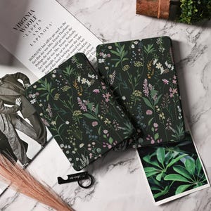 May include: Two notebooks with a dark green floral pattern featuring various colorful flowers. The notebooks are open. A black bottle opener and a book with text are also visible. The text reads 'Virginia Woolf and Lapinova'.
