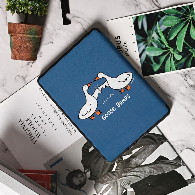 May include: A blue tablet case featuring a cartoon illustration of two white geese with orange feet and beaks, facing each other above wavy lines. The text "Goose Bumps" is below the geese. The case is on a marble surface.