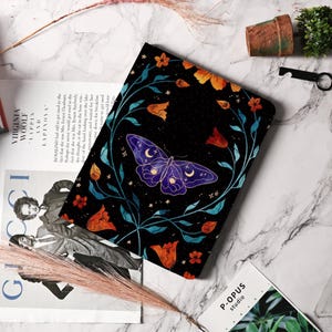 May include: A black notebook cover featuring a purple butterfly with moon and star details. The butterfly is surrounded by orange and red flowers and teal vines. The cover is on a marble surface.