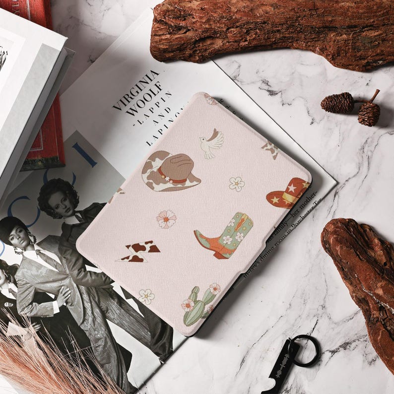 May include: A pink e-reader case with a western-themed design. The case features a cowboy hat, boots, cacti, and stars. The text "Virginia Woolf - Lappin and Lapin" is printed on the back of the case.