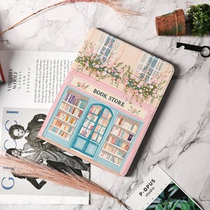 May include: A decorative box with a bookstore design. The box features a pastel pink and blue facade with the words "BOOK STORE". The design includes windows, floral accents, and shelves filled with books. The box is rectangular and has a hinged lid.