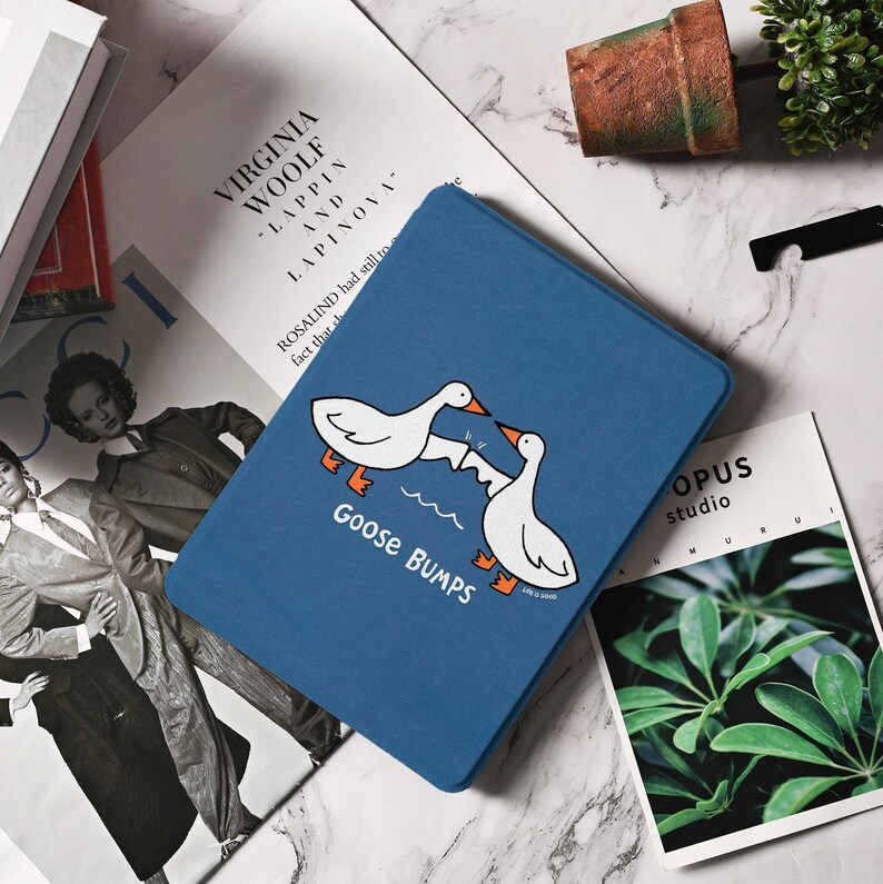 May include: A blue tablet case featuring a cartoon design of two white geese with orange feet and beaks, facing each other. The text "Goose Bumps" is below the geese. The case is on a marble surface with books and a plant.