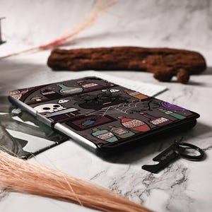 Pu&ograve; includere: Una custodia per e-reader illustrata in bianco e nero che presenta uno scaffale di pozioni con varie bottiglie, un teschio, una ragnatela e una pianta. La custodia ha un bordo nero e un portachiavi in metallo argentato.