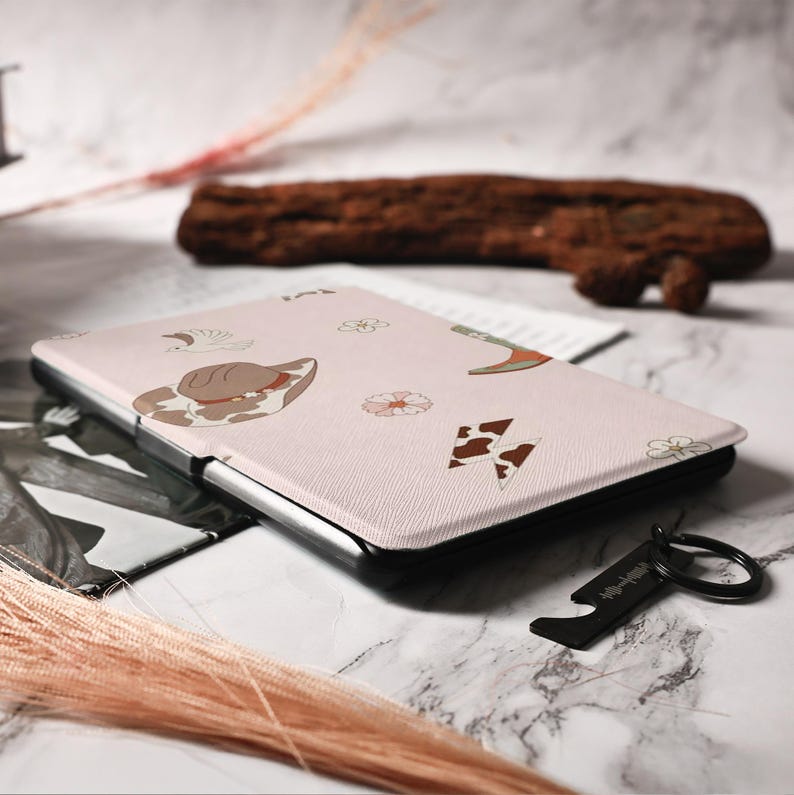 May include: A pink e-reader case with a cow print design and a variety of other patterns, including flowers and hats. The case is closed and lying on a table.