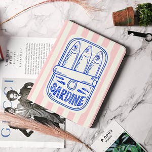 Könnte beinhalten: Tablet-Hülle mit rosa-weiß gestreiftem Muster. Sie zeigt eine blaue Illustration einer Sardinenbüchse mit drei Fischen und dem Wort "SARDINE". Die Hülle liegt auf einer Marmoroberfläche.