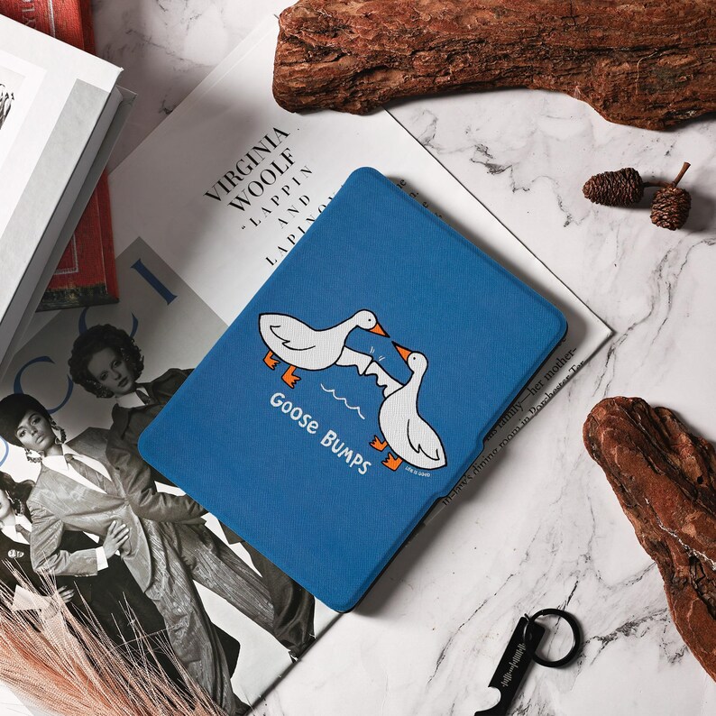 May include: A blue tablet case with a cartoon illustration of two white geese facing each other above the text "Goose Bumps". The case is resting on a magazine with the title "Virginia Woolf - Lappin and Lapinova".
