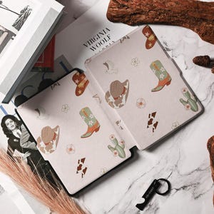 May include: A pink and white e-reader case with a cowboy boot, hat, and cactus pattern. The case has the text "Virginia Woolf" printed on the front.