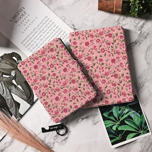 May include: Two pink floral patterned Kindle e-reader cases. The cases feature a delicate floral design with pink flowers and green leaves on a light pink background.