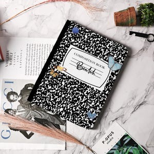 Puede incluir: Un cuaderno de composición blanco y negro con un patrón de cuadros blanco y negro. El cuaderno tiene una etiqueta blanca con el texto "COMPOSITION BOOK" y el nombre "Beulah" escrito en cursiva. El cuaderno tiene una pegatina de mariposa azul y una pegatina de planeta dorado y azul.