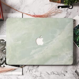 Puede incluir: Una funda para portátil con diseño de mármol verde claro. Tiene un recorte para el logotipo de Apple. La estética general es una mezcla de piedra natural y tecnología moderna.