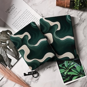 May include: Two dark green notebooks with a flowing white and light green pattern. The notebooks are on a marble surface, next to a black bottle opener and a magazine with the text "VIRGINIA WOOLF AND LAPINOVA".