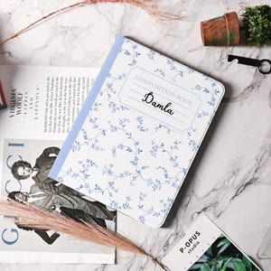 Puede incluir: Un cuaderno de composición blanco con un patrón floral azul y el texto "COMPOSITION BOOK" y "Damla" en la portada.