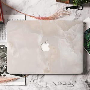 May include: A laptop case with a marble-like design in shades of white and beige. The case has a cutout for the Apple logo. The overall aesthetic is minimalist and elegant, suitable for protecting a laptop.