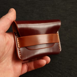 Op de afbeelding: Een kleine, bordeauxrode leren portemonnee met een contrasterende bruine band. De portemonnee wordt in een hand gehouden en toont de gladde, glanzende afwerking van het leer. De randen zijn netjes gestikt met een lichtgekleurde draad.