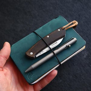 Könnte beinhalten: Ein kleines, dunkelgrünes Ledernotizbuch mit einem braunen Taschenmesser und einem silbernen Stift, die mit einem braunen Lederband befestigt sind.