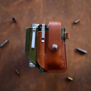 May include: Two leather sheaths, one olive green and one brown, with silver metal clips. The brown sheath has a flap secured by a silver snap. The sheaths are on a brown leather surface, with several small metal objects scattered around.