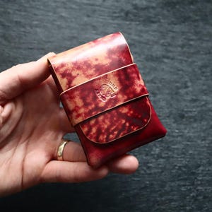Può includere: Un porta carte in pelle rettangolare con una finitura maculata rossa e marrone chiaro. L'accessorio include una cinghia orizzontale al centro con un logo circolare in rilievo. La pelle presenta una texture marmorizzata con toni cremisi.