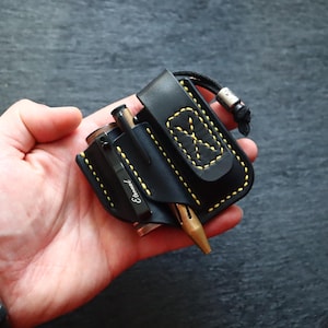 Puede incluir: Organizador EDC de cuero negro con costuras amarillas, que contiene un bolígrafo y una pequeña linterna. El organizador tiene un clip para cinturón y un cordón. El bolígrafo tiene la palabra "Eternal".