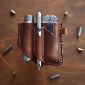 Puede incluir: Una funda de cuero marrón para herramientas multiusos con tres ranuras. La funda tiene un borde cosido y una pequeña correa de cuero. La funda contiene un bolígrafo, una herramienta multiusos y una linterna.