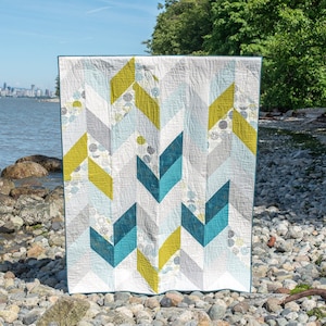 May include: A contemporary quilt featuring a chevron design in teal, grey, and chartreuse. The quilt is shown outdoors near a rocky beach with a city skyline in the background. The quilt's pattern incorporates geometric shapes.