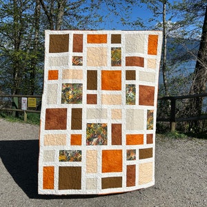 May include: A patchwork quilt with a geometric design featuring squares and rectangles in shades of brown, orange, and cream. The quilt is made with a variety of fabrics, including a floral print.