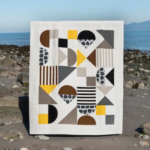 May include: A geometric quilt with a white background and various shapes in shades of brown, grey, black, and yellow. The quilt is made of different fabrics with patterns like stripes, polka dots, and solid colours.