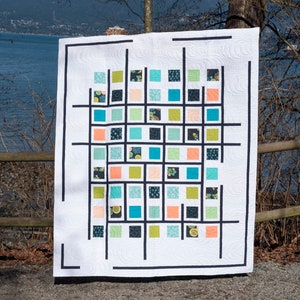 May include: A white quilt with a geometric pattern of black lines and colorful squares. The squares are in shades of blue, green, orange, and yellow. The quilt is displayed against a backdrop of a body of water and trees.