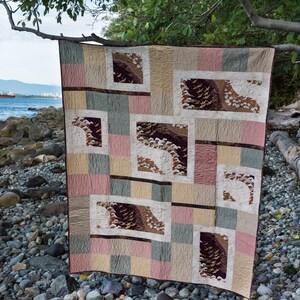 May include: A patchwork quilt with a geometric pattern of squares and rectangles. The quilt is made of various fabrics in shades of brown, beige, and cream. The quilt is hanging on a tree branch over a rocky beach.