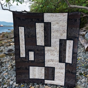 May include: A black and white geometric patterned quilt with a modern design. The quilt is made of fabric with a textured surface. The quilt is lying on a bed of rocks.