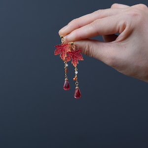 Op de afbeelding: Een paar goudkleurige bungelende oorbellen met doorschijnende rode esdoornbladbedels. Elke oorbel bevat een fijne gouden ketting met kleine witte en oranje kralen, eindigend in een gladde, druppelvormige rode glazen hanger.