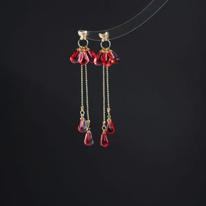 Puede incluir: Un par de pendientes colgantes con un diseño floral. Los pendientes presentan colgantes rojos en forma de pétalo y pequeños detalles de mariposa dorada. Delicadas cadenas doradas conectan los colgantes con las partes superiores de mariposa. El fondo es negro liso.