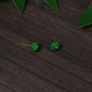 Puede incluir: Un par de pendientes de jade verde con postes y detalles dorados. Los pendientes son redondos con un diseño tallado, sobre un fondo de vetas de madera oscura. Una joya clásica.