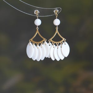 Puede incluir: Un par de pendientes dorados con detalles blancos. Cada pendiente presenta una cuenta blanca en la parte superior, un elemento dorado en forma de abanico y múltiples colgantes blancos de forma ovalada. Los pendientes se muestran sobre un fondo borroso verde y marrón.