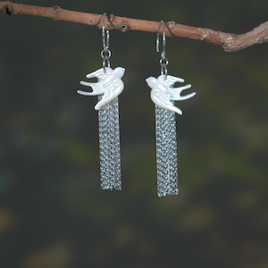 May include: A pair of dangling earrings featuring white bird charms with silver chain fringe. The earrings are suspended from silver hooks and are displayed against a blurred green and brown background, attached to a small branch.