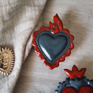 Peut inclure: Trois objets décoratifs en forme de cœur. La pièce centrale est un cœur gris avec une bordure festonnée et des flammes rouges. Deux autres cœurs sont visibles, un doré et un rouge avec une bordure grise. Les objets sont sur une surface en bois.