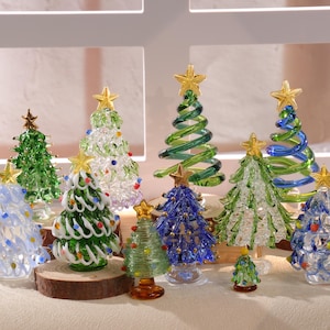 May include: A collection of colorful glass Christmas trees in various sizes and designs. Each tree features a gold star on top and is decorated with clear, green, blue, and white glass. Some trees are on wooden bases.