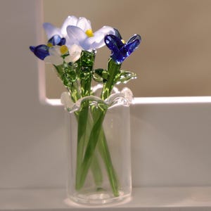 May include: A glass vase holding a bouquet of glass flowers. The flowers are in shades of blue, white, and yellow, with green stems and leaves. The vase is clear glass with a ruffled rim, and the flowers are arranged in a natural, organic style.