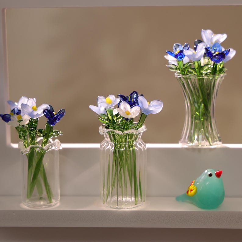 May include: Glass floral arrangements in clear vases. The flowers have blue, white, and yellow petals. A small, turquoise glass bird with a red beak and yellow accents sits nearby. The vases are on a white shelf.